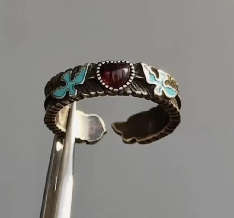 Vintage enamel ring with a garnet stone held in tweezers, shot on a phone against a grey background