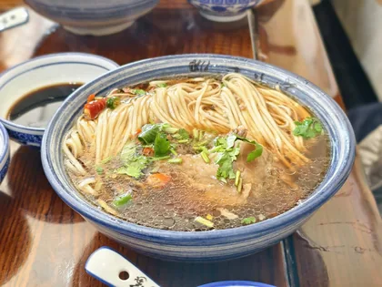 A bowl of noodles shot on a phone at a restaurant table under warm light, with other bowls around