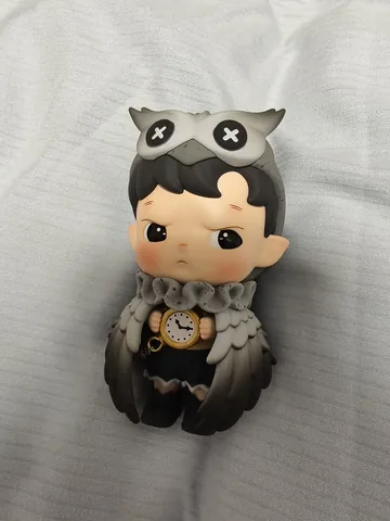 A grey owl-hooded art toy shot on a phone, on white fabric